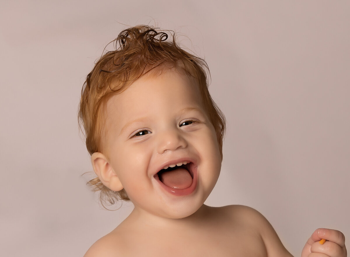 Baby boy with red hair is laughing at the camera during is first birthday photoshoot in brooklyn, ny. Close up image of his face. Captured by premier Brooklyn NY family photographer Chaya Bornstein Photography.