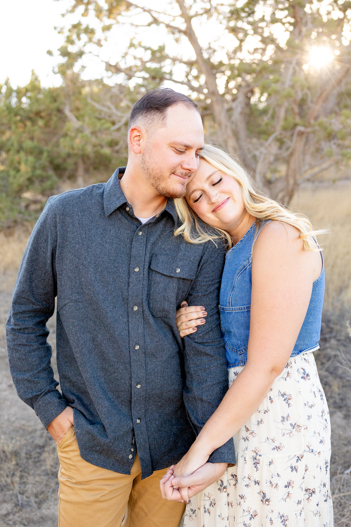 A woman leans her head on her partner’s shoulder as they hold hands, soaking in a peaceful moment together. The golden light filters through the trees, enhancing the romantic feel.