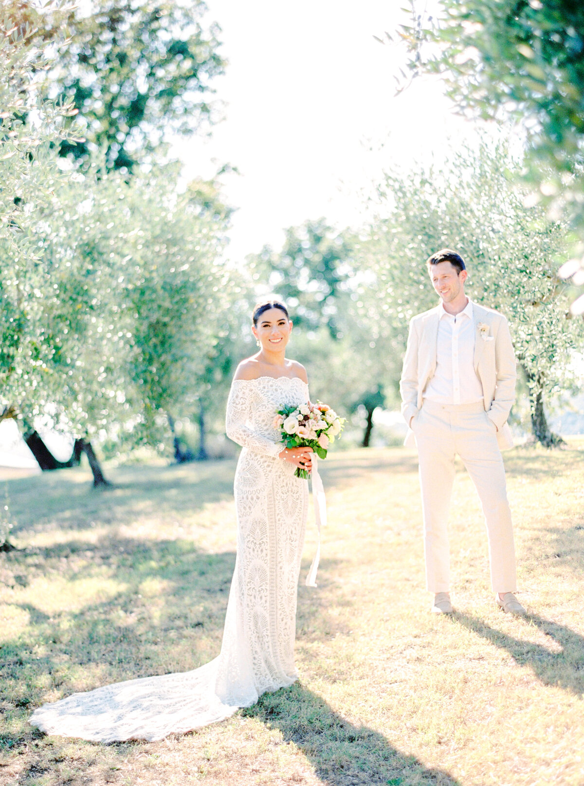Film photograph of bride in all lace long sleeved off the shoulder wedding dress standing in the sun while the groom in a soft light tan wedding suite looks at her adoringly photographed by Italy wedding photographer at Villa Montanare Tuscany wedding