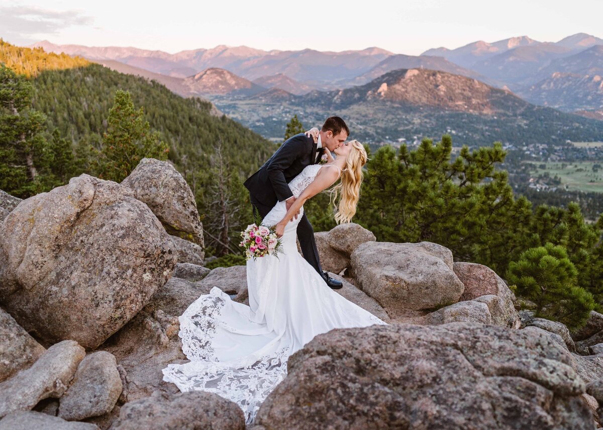  Groom dips and kisses bride atop rocky cliffs with panoramic sunset views over Estes Park, Colorado.