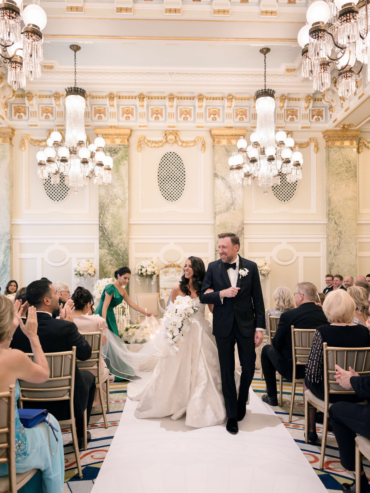 A couple walks down the aisle in an elegant wedding venue adorned with chandeliers and ornate decor. The bride wears a flowing white gown with a long train and holds a bouquet, while the groom is in a black tuxedo. Guests sit on either side, capturing the moment.