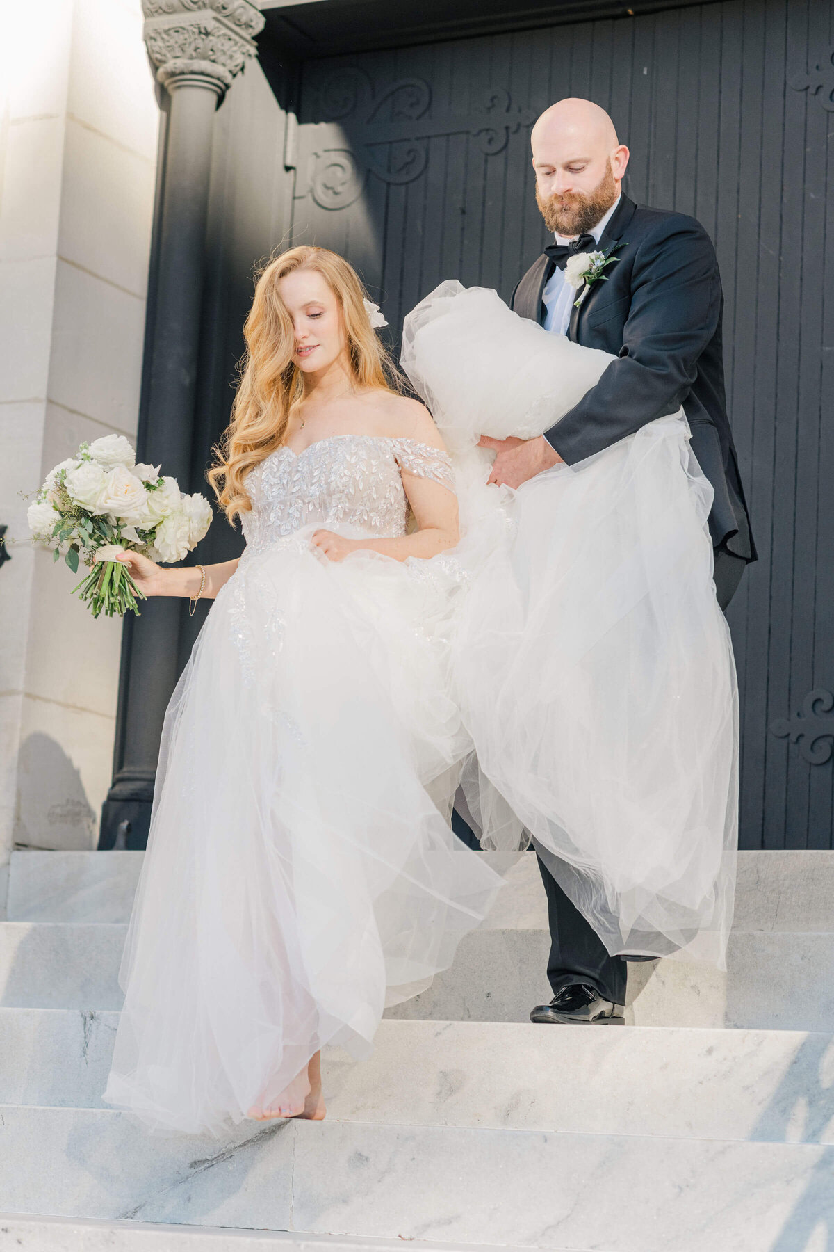 Bride walking down steps with bouquet while groom holds up wedding dress