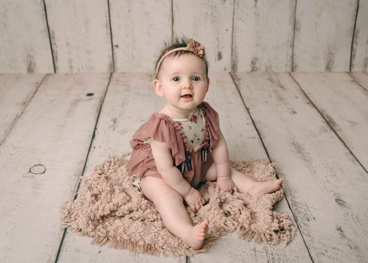 Six Month Baby Girl Sitting on Pink blanket Dressed in Pink Floral Romper with matching bow headband