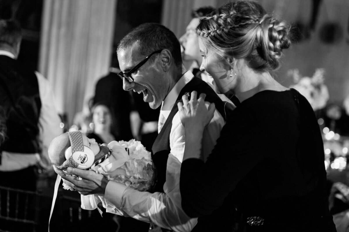 Candid wedding reception moment at The Thompson Hotel with the couple dancing while holding their newborn baby wearing earmuffs.