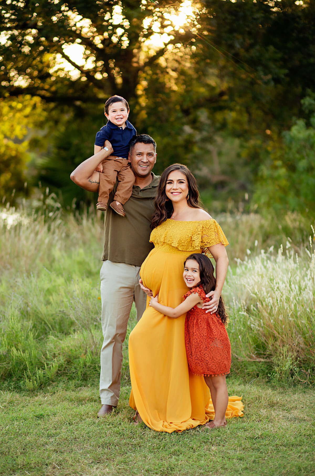 pregnant mother in yelow dress poses with her family while daughter hugs belly and dad holds son on his shoulder