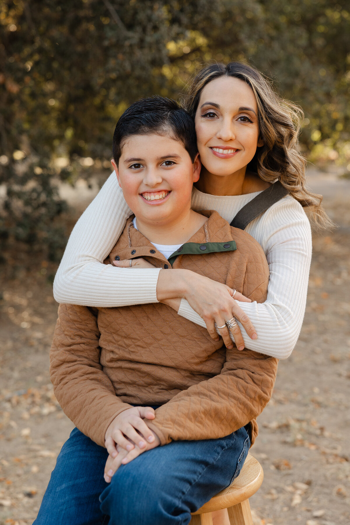 Mother hugging son from behind while he sits on wooden chair outdoors. Photographed by Yucaipa family photographer Kaitlyn Dawn Photography.