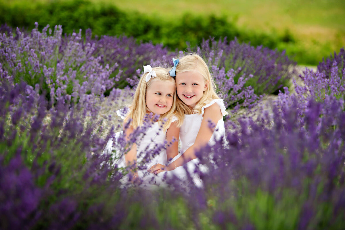two blond-haired sister sitting on the ground with purple lavender all around them for their family photos.  Award-winning photo.