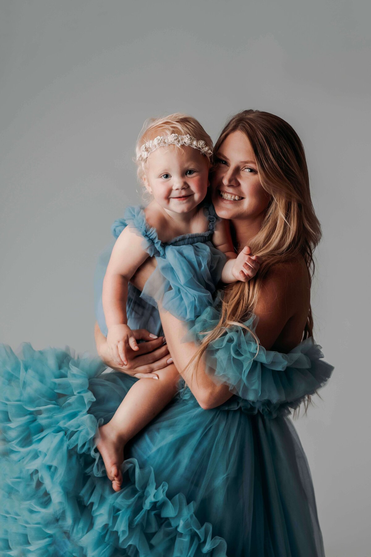 A close-up photo captures a young mom and her toddler daughter, both wearing matching blue tulle gowns, cheek to cheek and smiling at the camera. Their joyful expressions highlight their close bond, with the child wearing a white floral headband