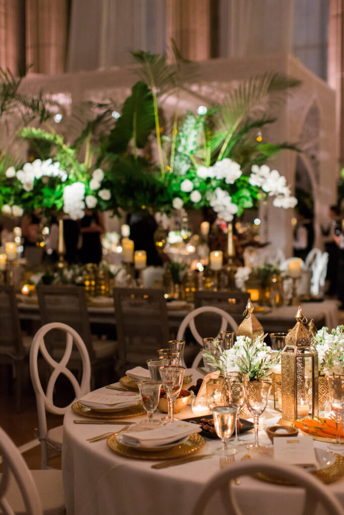 At the exquisite Mellon Auditorium in Washington D.C., photographer Sarah Bradshaw captured the essence of luxury with European flair. The photo showcases an elegantly set dining table adorned with white and gold tableware, accented by lush green tropical leaves and white flowers as the centerpiece. The warm lighting from lanterns and candles enhances the ambiance. In the background, additional decorated tables and greenery further elevate the sophisticated setting.