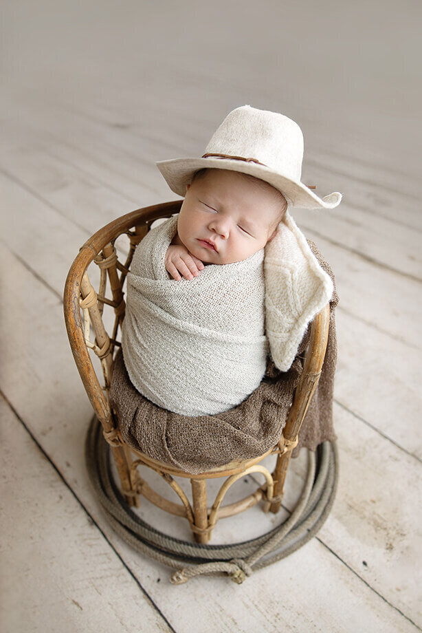 newborn boy is swaddled in a cream wrap, wearing a matching cowboy hat and posed in a wicker chair and decorated with a lasso.