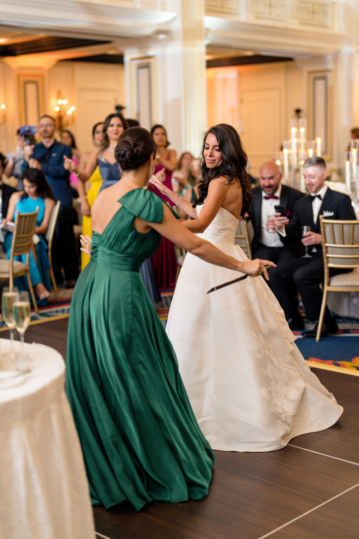 Two women dance at a formal event. One wears a white strapless gown, and the other a green dress. Guests, some seated and some standing, watch and capture the moment with phones. The room is elegantly decorated with candles and chandeliers.