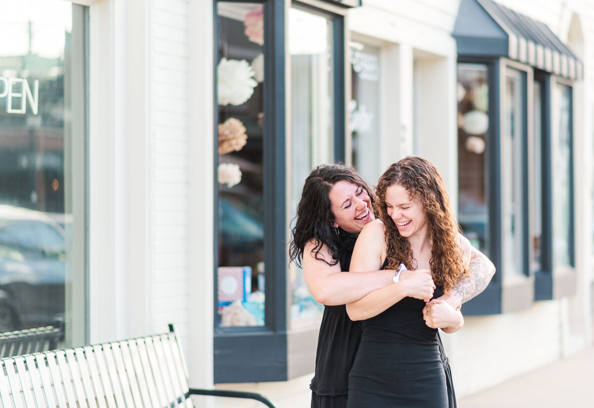 Two women in black dresses for an engagement session captured by denver wedding photographer hug and smile towards each other