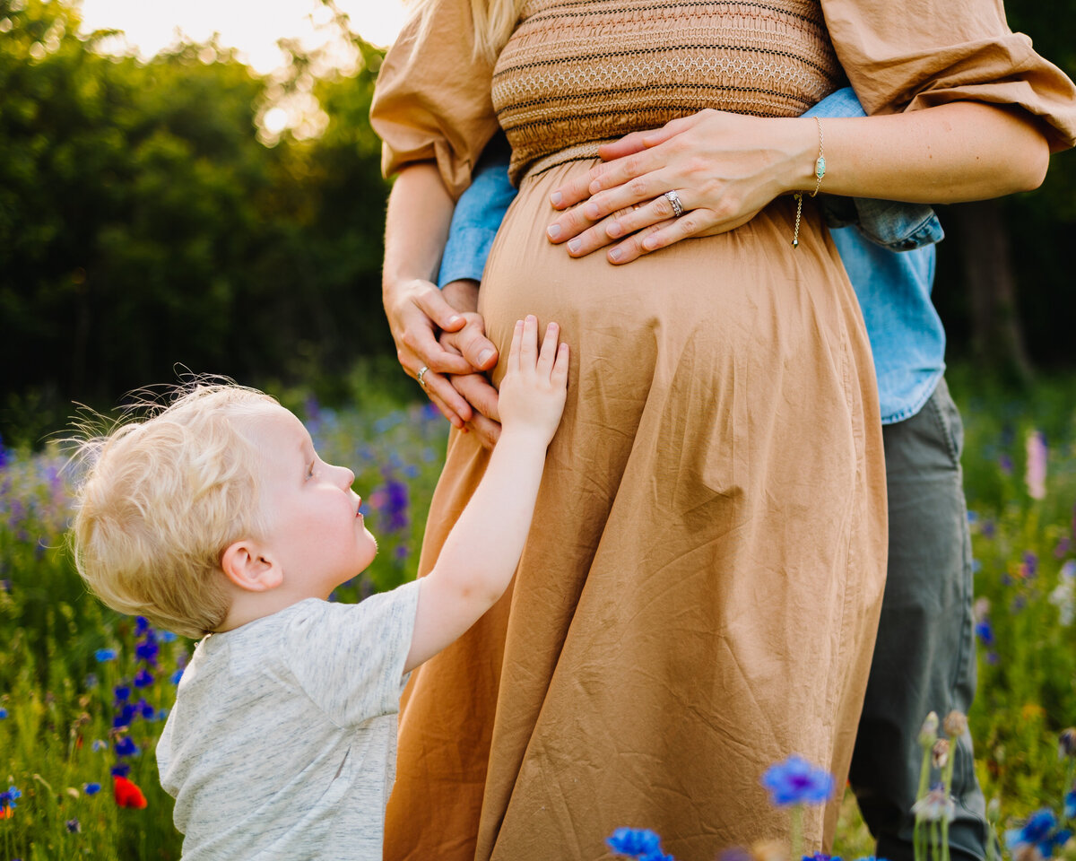 In this heartwarming family maternity photo, a pregnant woman in a long brown dress is surrounded by her loved ones in a garden. The child gazes at her belly with curiosity, while the man in a blue shirt embraces her lovingly.