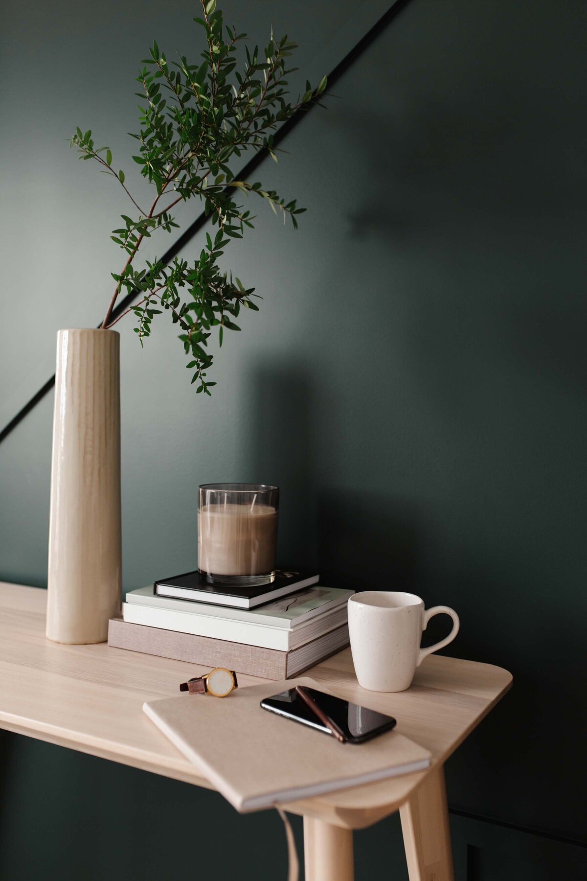 dark green walls with light oak desk top, thick books with candle, white mug, journal, watch, iphone