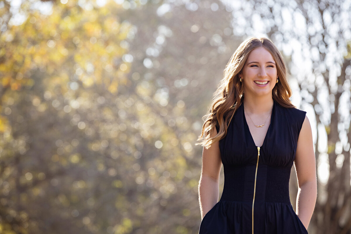 High school senior in black dress with golden trees and gravel path in Charlotte-area nature preserve