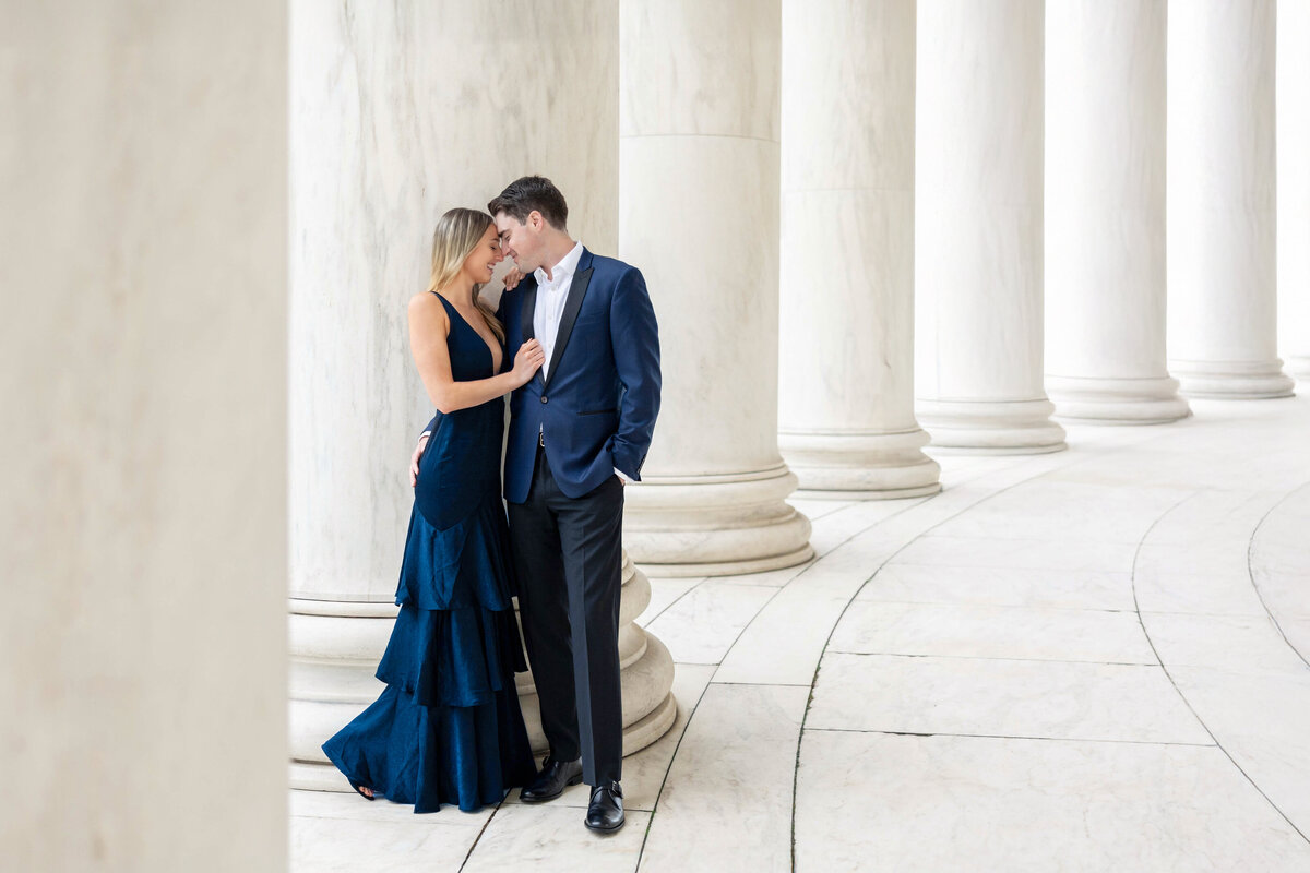 A couple dressed elegantly, with the woman in a navy gown and the man in a suit, stand close together against a backdrop of large white marble columns. They are gazing at each other and smiling, creating a romantic atmosphere.