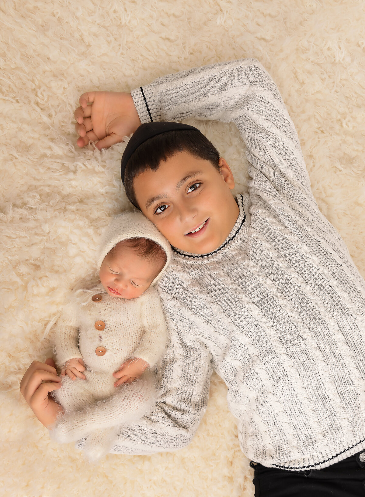 Big brother poses with his new baby brother for a brooklyn newborn photoshoot. Aerial image. Big brother is laying on a cream fluffy rug wiht baby's head resting atop of his shoulder. Big brother is smiling at the camera as baby sleeps. Captured by top Brooklyn newborn photographer Chaya Bornstein