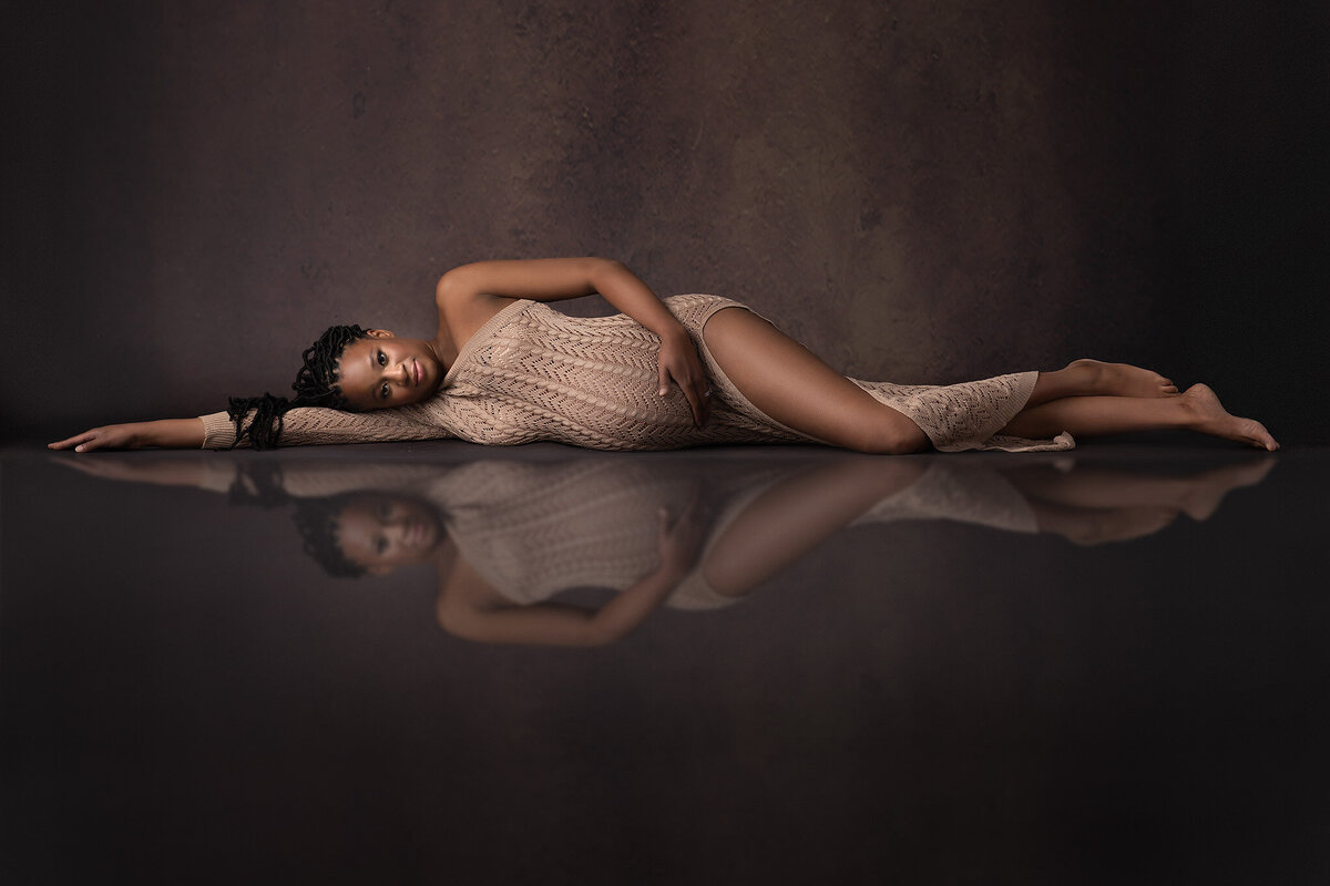 A pregnant woman reclines gracefully in a form-fitting knit gown, her reflection mirrored beneath her. Captured by DFW maternity photographer Lindsay Walden, this elegant maternity photograph showcases warmth and tranquility.