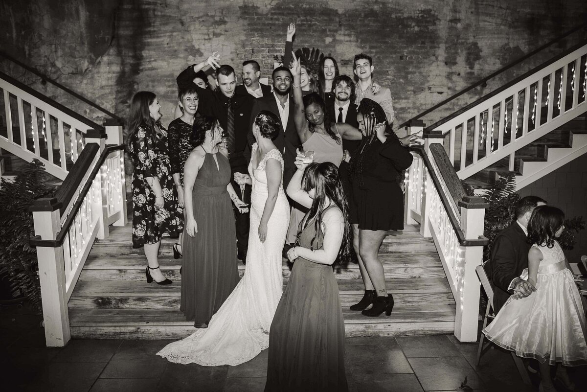A bride and groom stand on stairs surrounded by joyful friends and family, some posing, smiling, or laughing. The group is dressed formally. The setting appears festive, with string lights on the railings. The mood is celebratory.