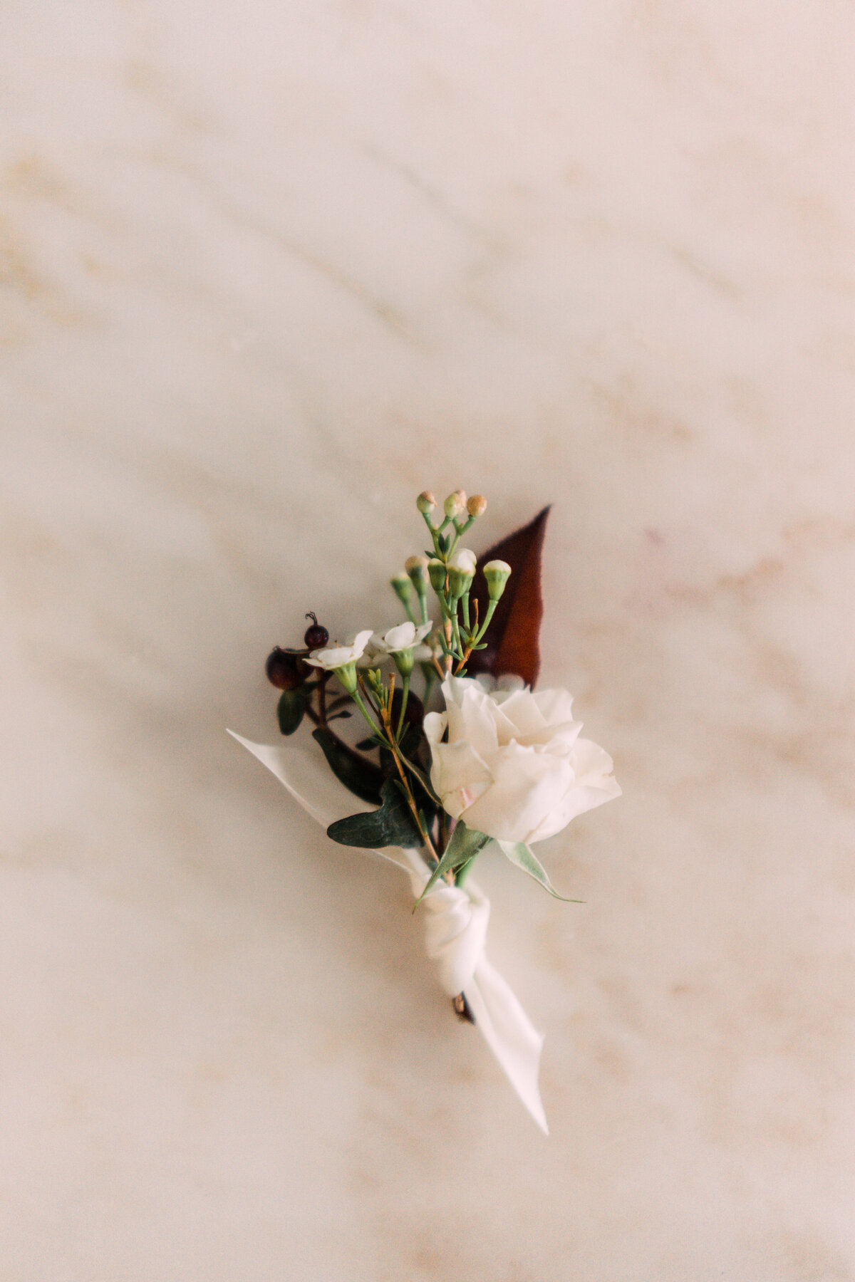 Boutonniere with burgundy hypericum berries & foliage, white spray rose & waxflower, & hand-tied ivory ribbon on marble table