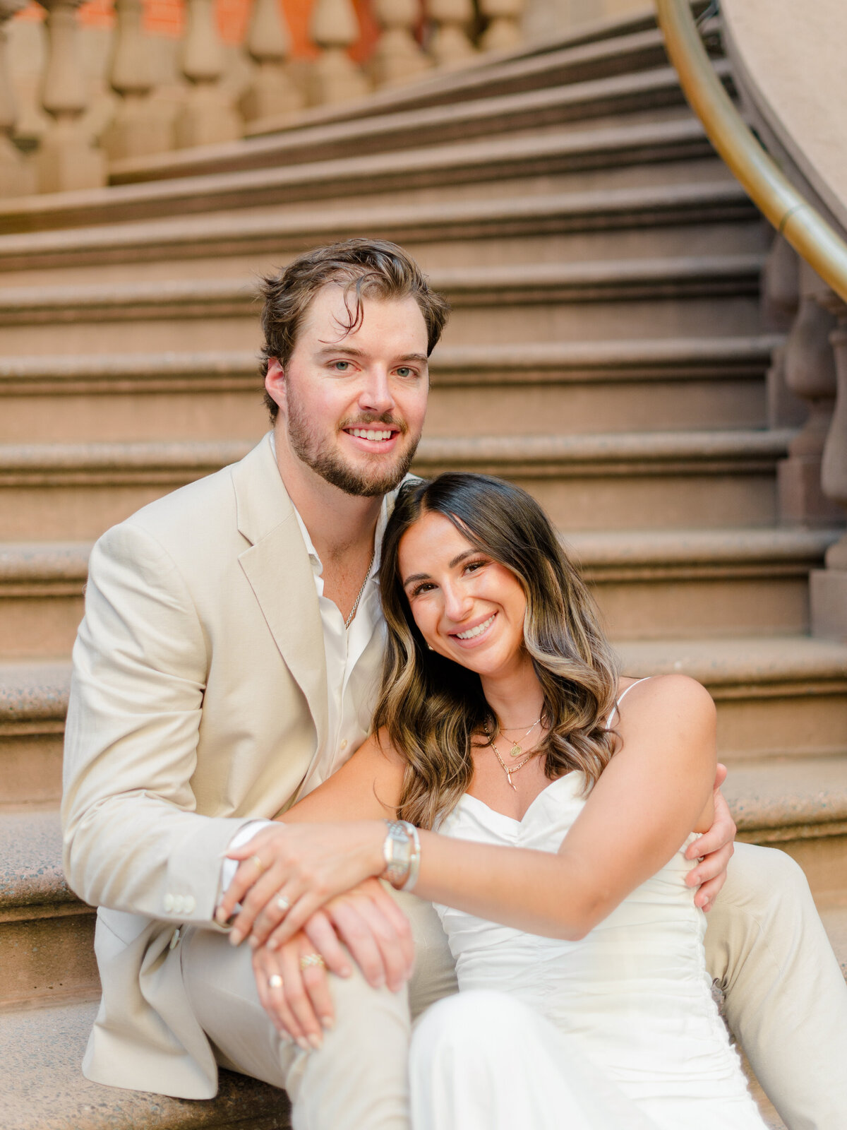 A couple sits closely on the iconic staircase outside The Union League of Philadelphia, smiling warmly at the camera. Dressed in a light suit and a white dress, they exude effortless elegance and joy. Captured by Elizabeth Kane Photography, a Luxury Philadelphia Wedding Photographer.
