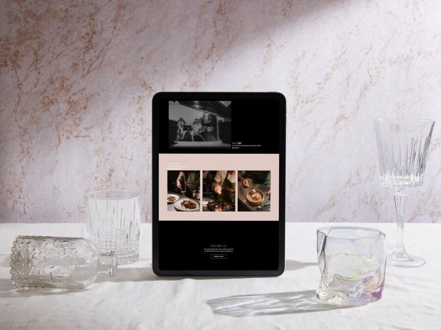 An iPad displaying the home page page of restaurant saels website, showcasing the food gallery section. The iPad is set on a white surface with crystal glassware nearby, creating an elegant and refined presentation.