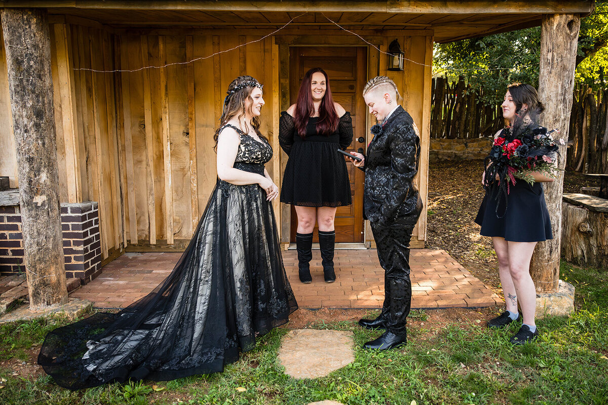 Two queer marriers stand in front of one another, as one bride wearing a floral suit reads their vows written on their phone. A friend of theirs stands in the middle to act as their officiant for the day of their ceremony while another friend stands off to the side holding a bouquet in a backyard of an Airbnb they rented.