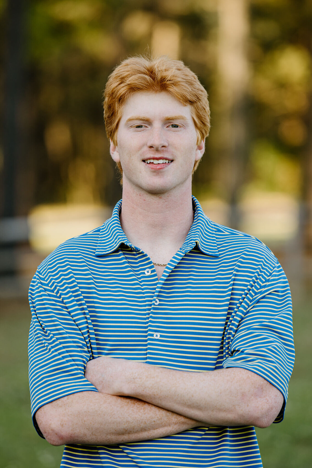 senior portrait of male graduate wearing blue and yellow striped polo shirt with arms crossed while standing in an East TX field