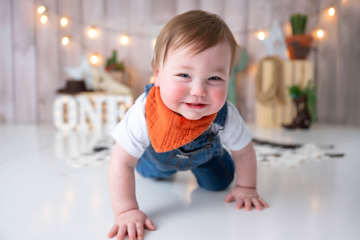 Smiling baby crawling away from a cowboy-themed backdrop decorated with various western props.