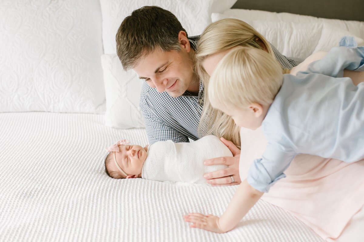 young family in northern virginia lays on bed during newborn photography session looking at new baby