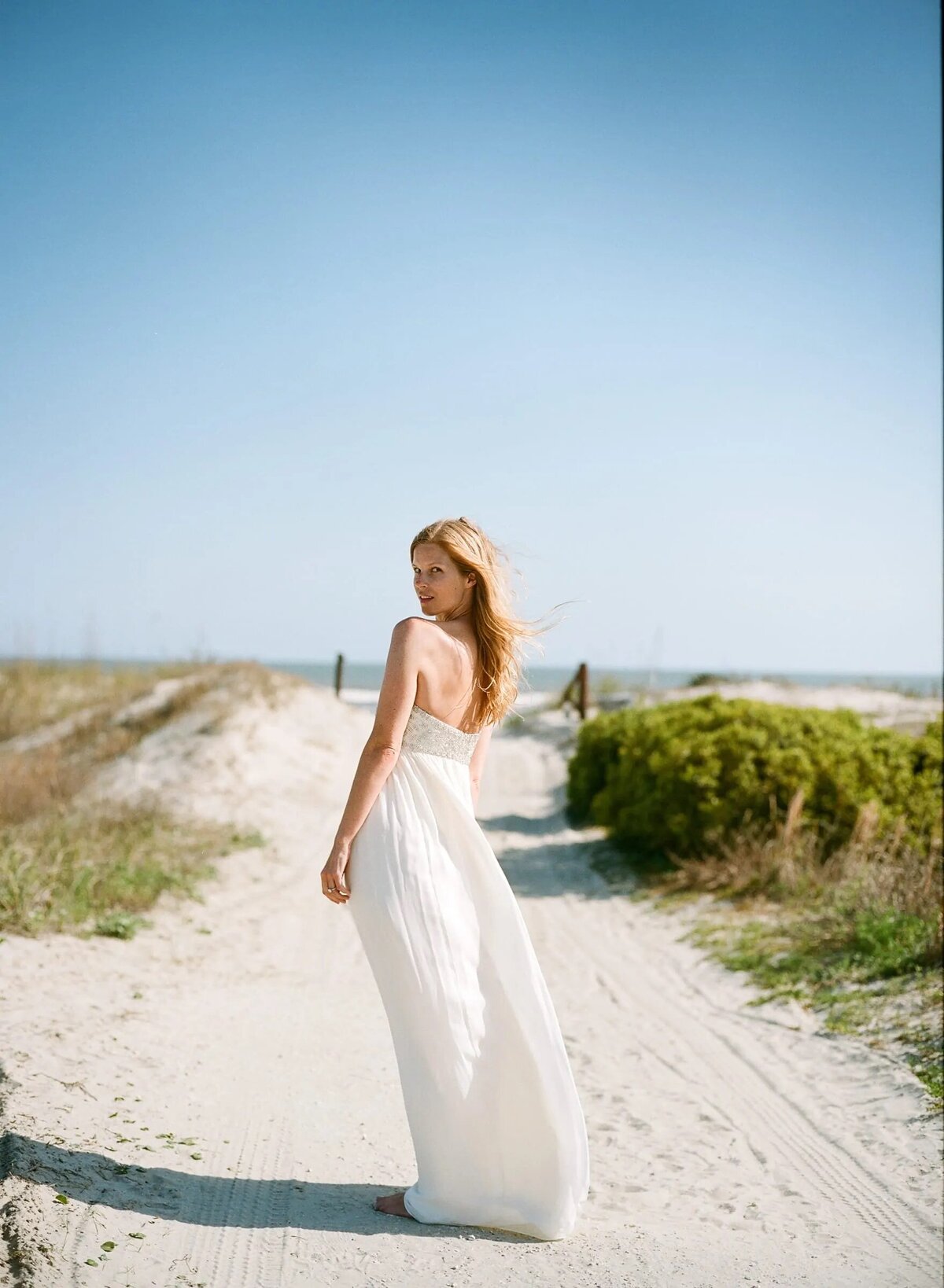 A bride in a flowing white gown looks back over her shoulder on a sandy beach path, with the sea in the distance, embodying a serene bridal moment.