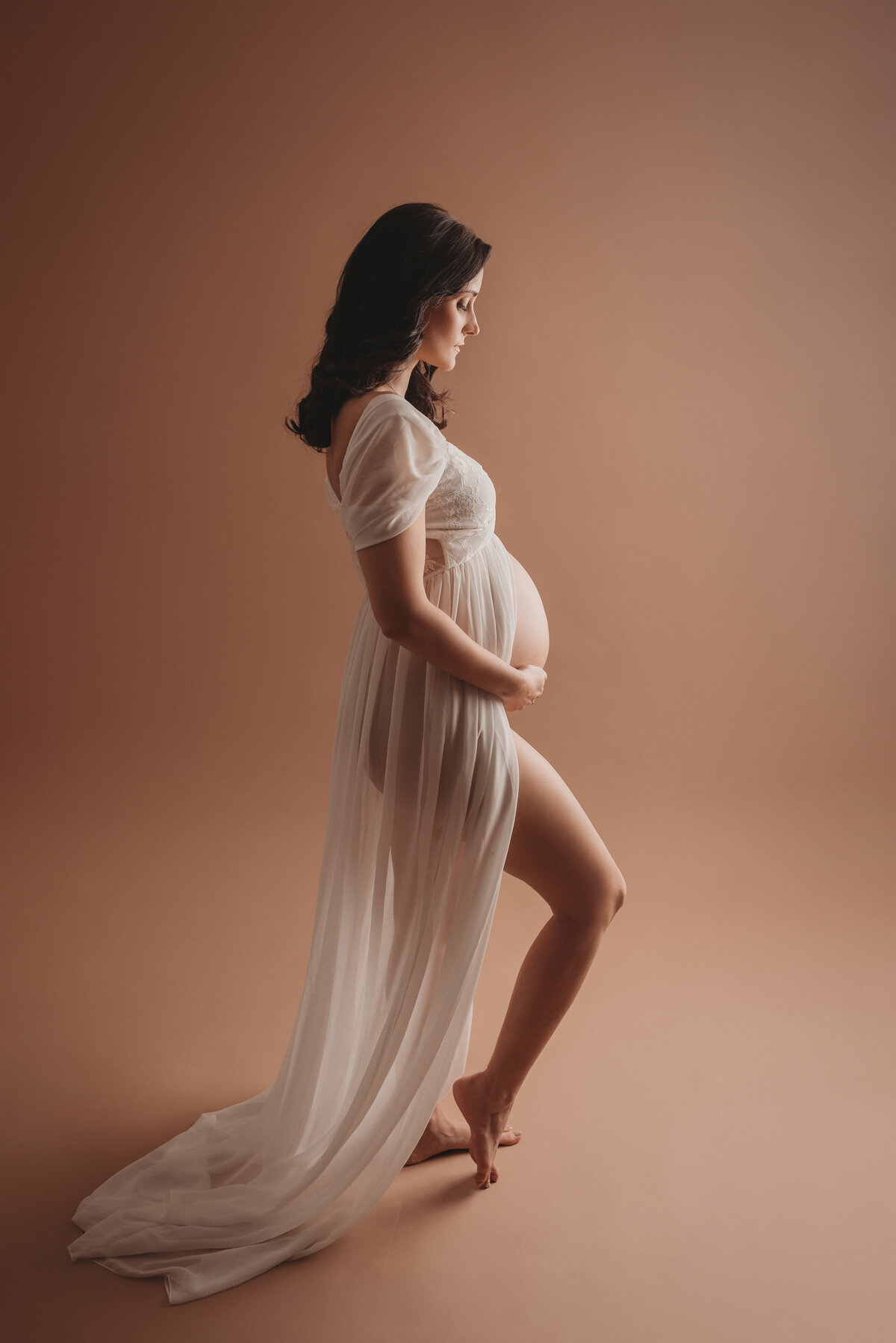 Pregnancy portrait on tan backdrop with 36 week pregnant woman holding baby bump looking at baby bump and wearing sheer white maternity gown open on tummy