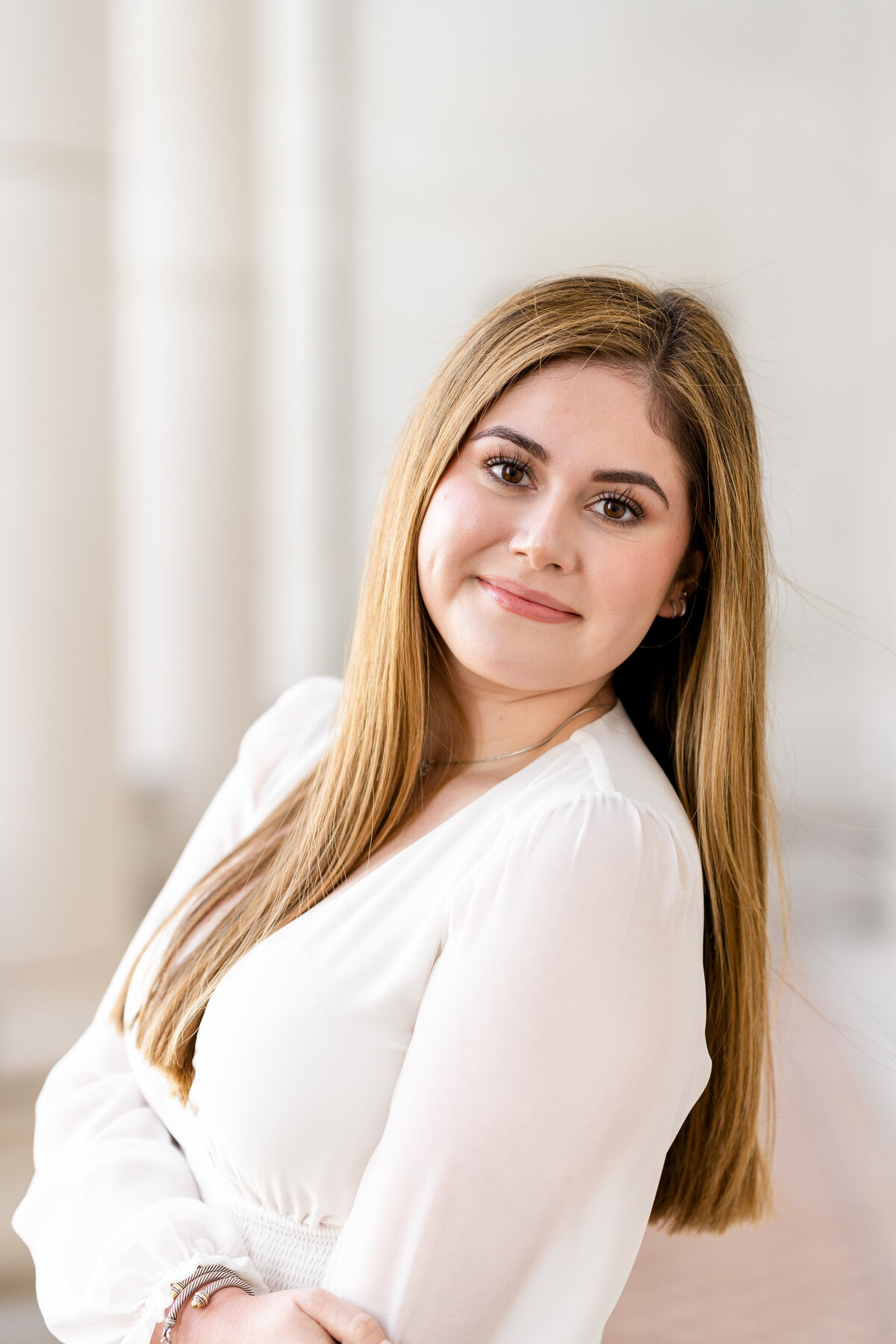 Texas A&M senior girl smiling at camera and looking over shoulder while hugging elbow and wearing white dress in columns of Administration Building