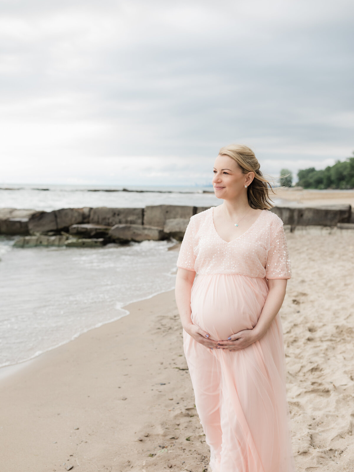pregnant mother in pink maternity gown holding baby bump on beach for maternity photoshoot cleveland maternity photographer