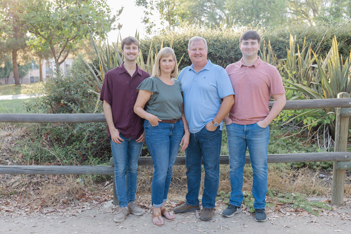 A family of four standing in front of a wooden park fence and lots of green trees and bushes, smiling at the camera with their hands in their jean pockets. Captured at Citrus Ranch Park in Tustin.