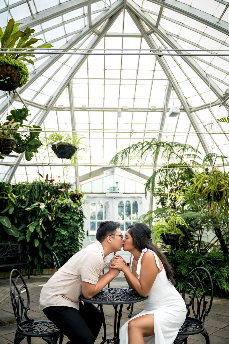 This romantic engagement photo captures a tender moment as the couple holds hands and kisses while seated at a black iron table inside the iconic Conservatory of Flowers in San Francisco. Surrounded by lush greenery and under the striking glass architecture, the couple radiates love and joy. The soft natural light filtering through the glass roof adds a serene and intimate vibe to the shot, making it a perfect backdrop for their engagement session.
