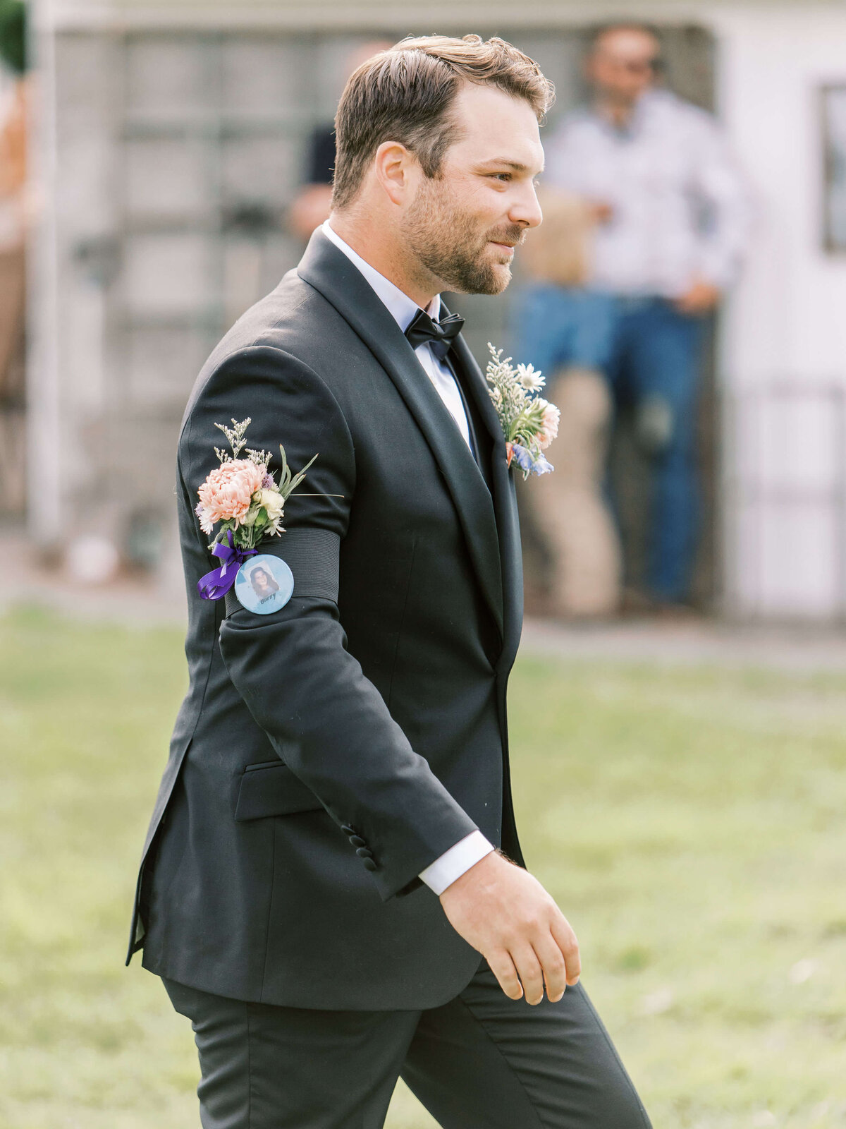A groom walks outdoors at a luxury backyard wedding, he wears a pin on his arm with a boutonnière of flowers in remembrance of a loved one.