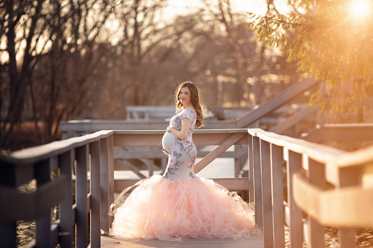 A mother to be in a blue floral print and pink tule maternity gown stands on a wooden dock at sunset