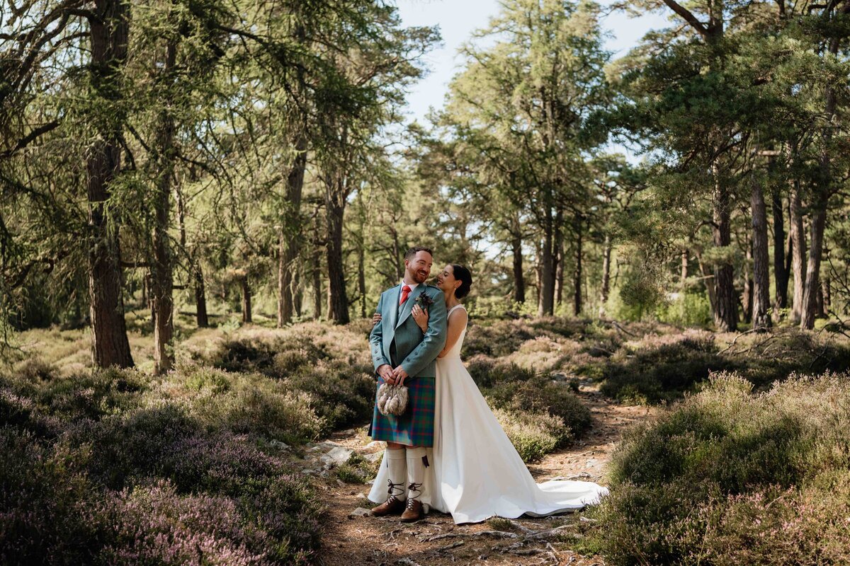 A wide photograph of a newly married couple wearing wedding attire. They stand in a clearing in the woods and heather blooms around them.
