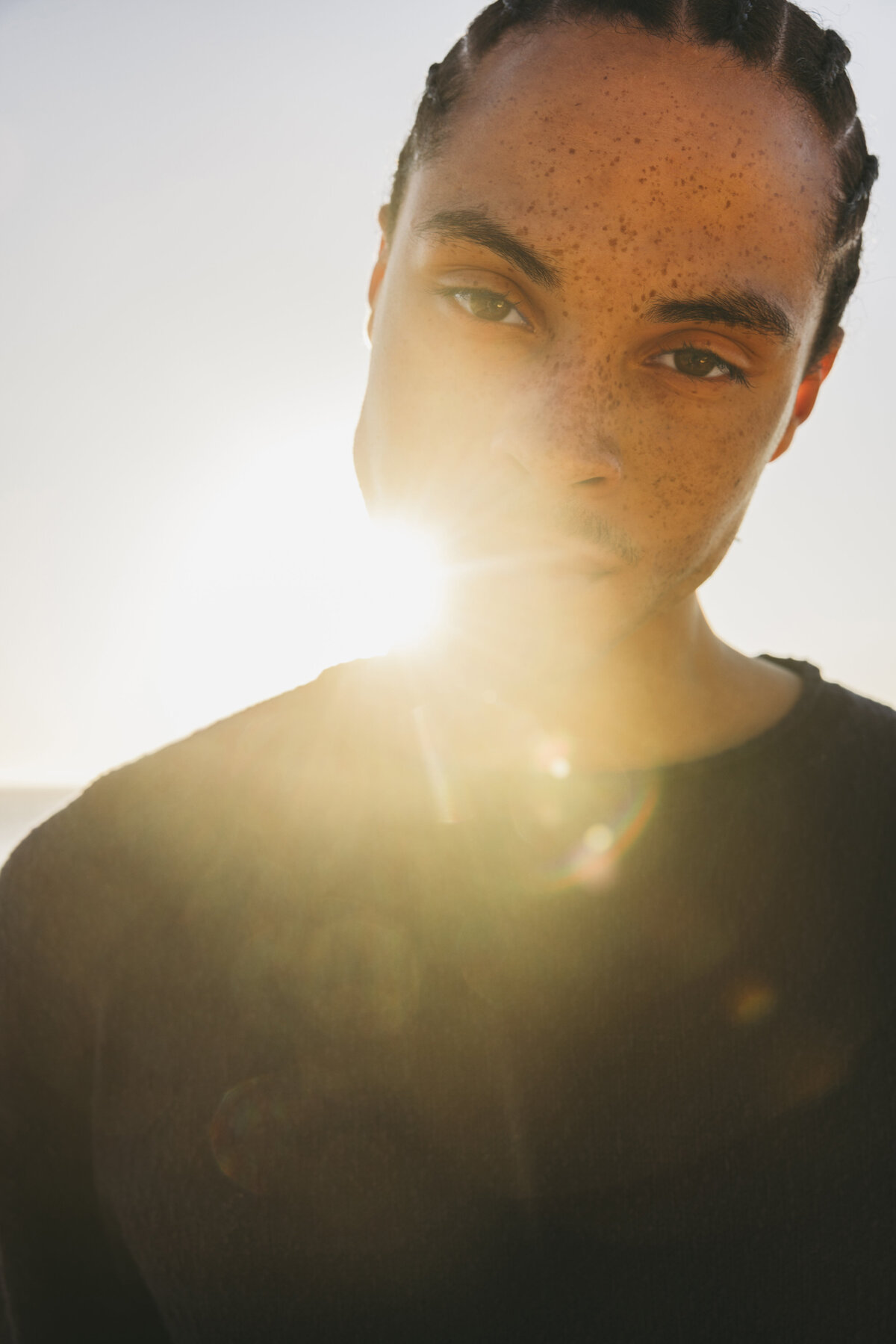 Close-up portrait of a male model with braids and freckles, backlit by the setting sun creating a strong lens flare effect in Cape Town.
