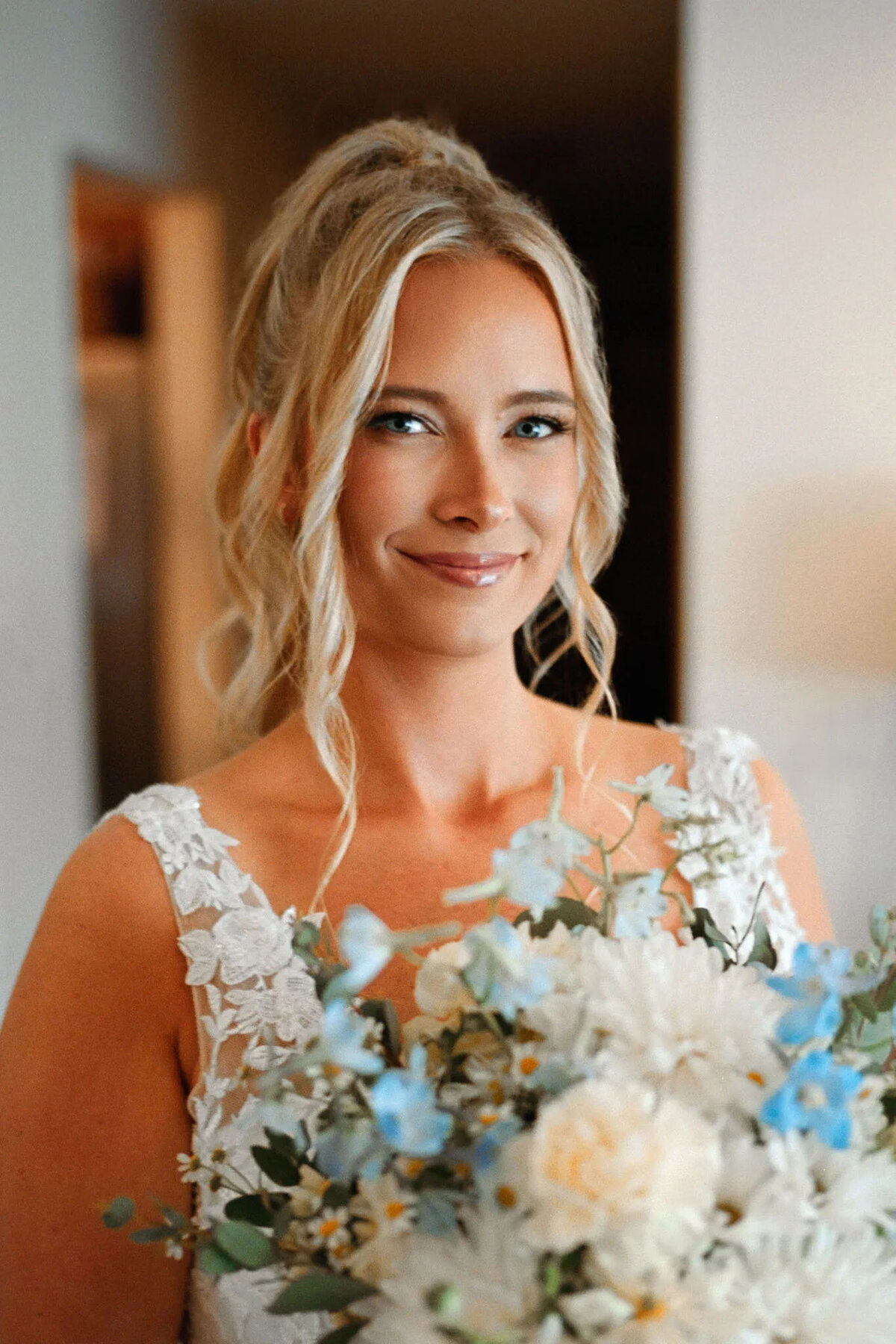 A woman with blonde hair in loose curls, wearing a white lace dress, holds a bouquet of white and light blue flowers and smiles softly indoors, captured beautifully by an NJ wedding photographer.