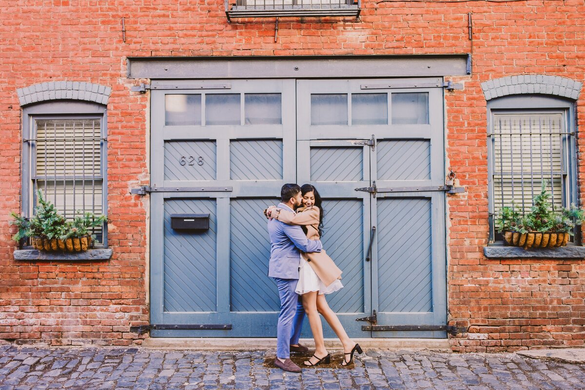 An engaged couple share a moment in front of a large door on a cobblestone street in hoboken nj in court street