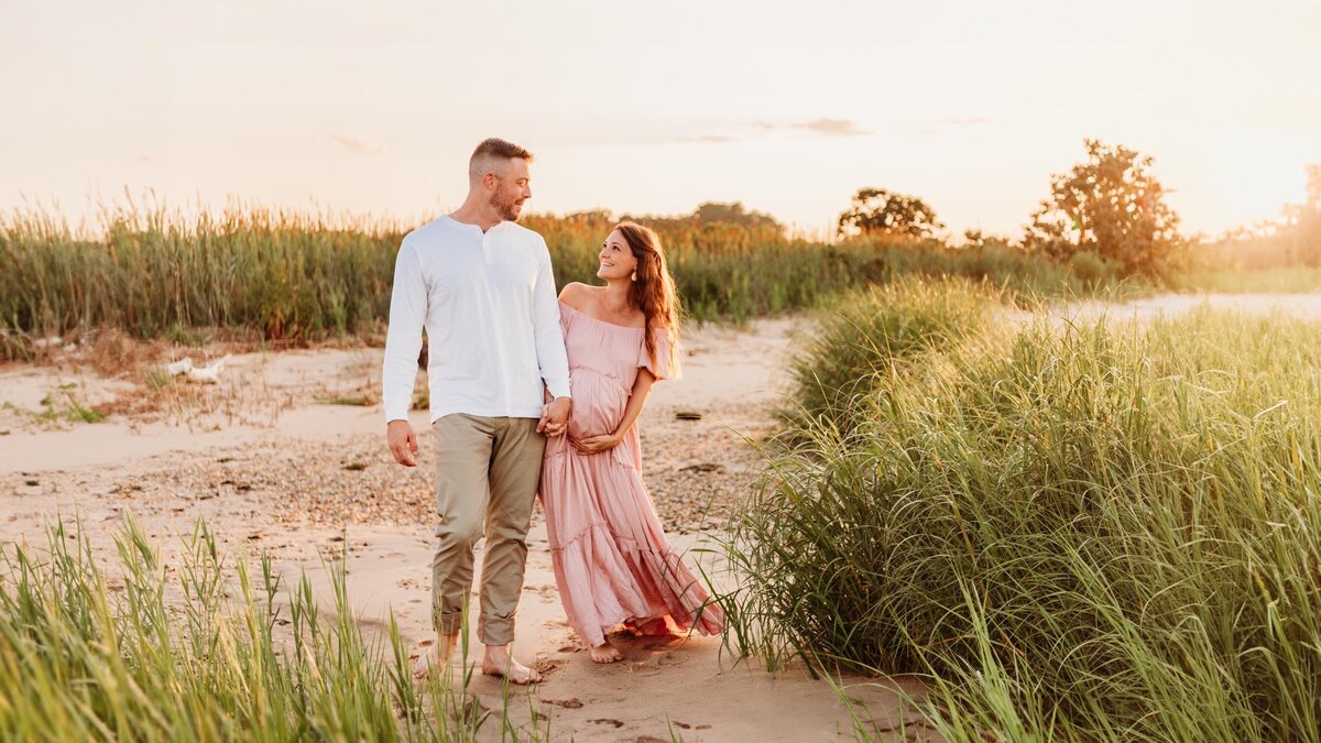 An expecting mom is cupping her baby bump, while holding her husband's hand. They are walking along the beach, smiling at one another with the golden sunlight behind them.  Photo taken by Delaware maternity photographer, Kristi