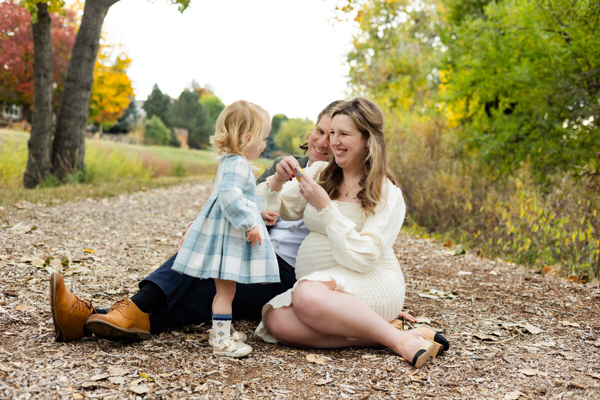 Mom and dad sit on the ground and smile at toddler daughter while feeding her a snack.
