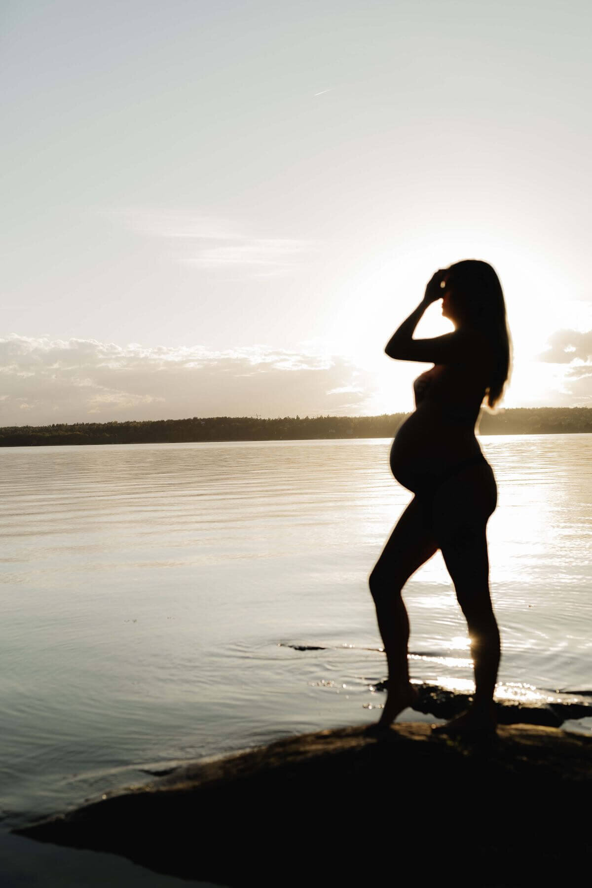 Bild på en gravid kvinna i siluett ståendes vid vattnet med ena handen i håret