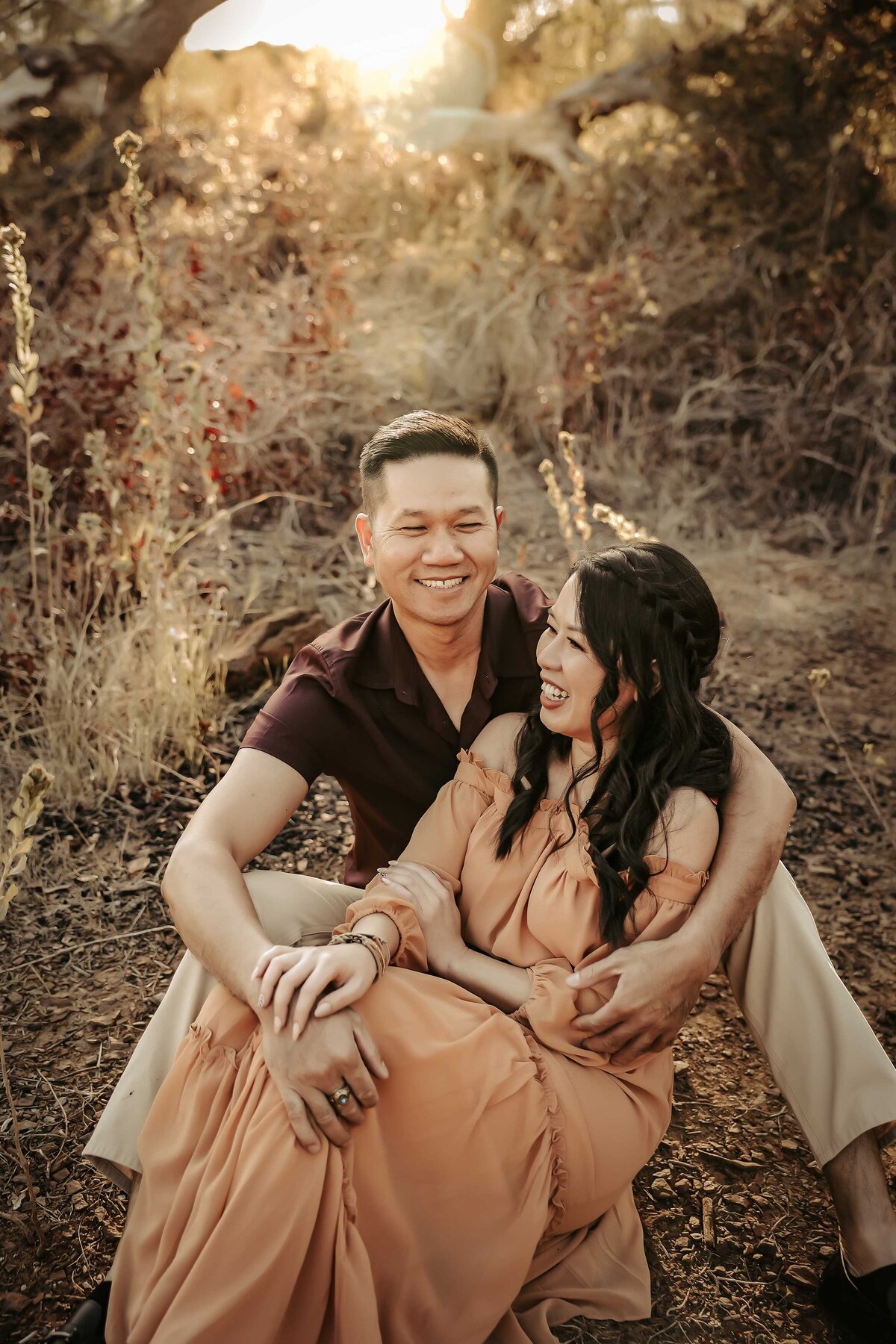 a man is sitting on the ground holding his wife in his arms while they laugh. She is wearing a light peach maxi dress and her hair is wavy.