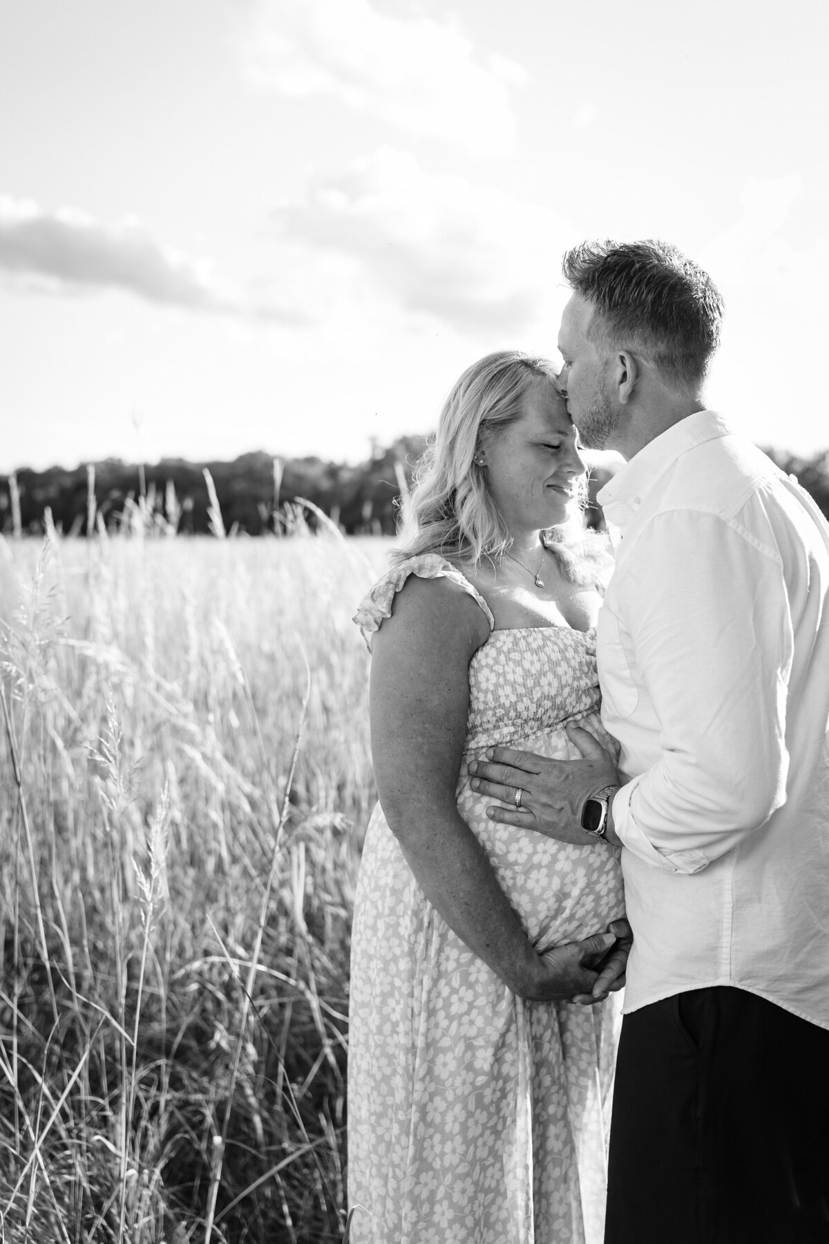 A black-and-white photo of a man gently kissing a pregnant woman's forehead as they stand in a grassy field, with the woman's hand on her belly.