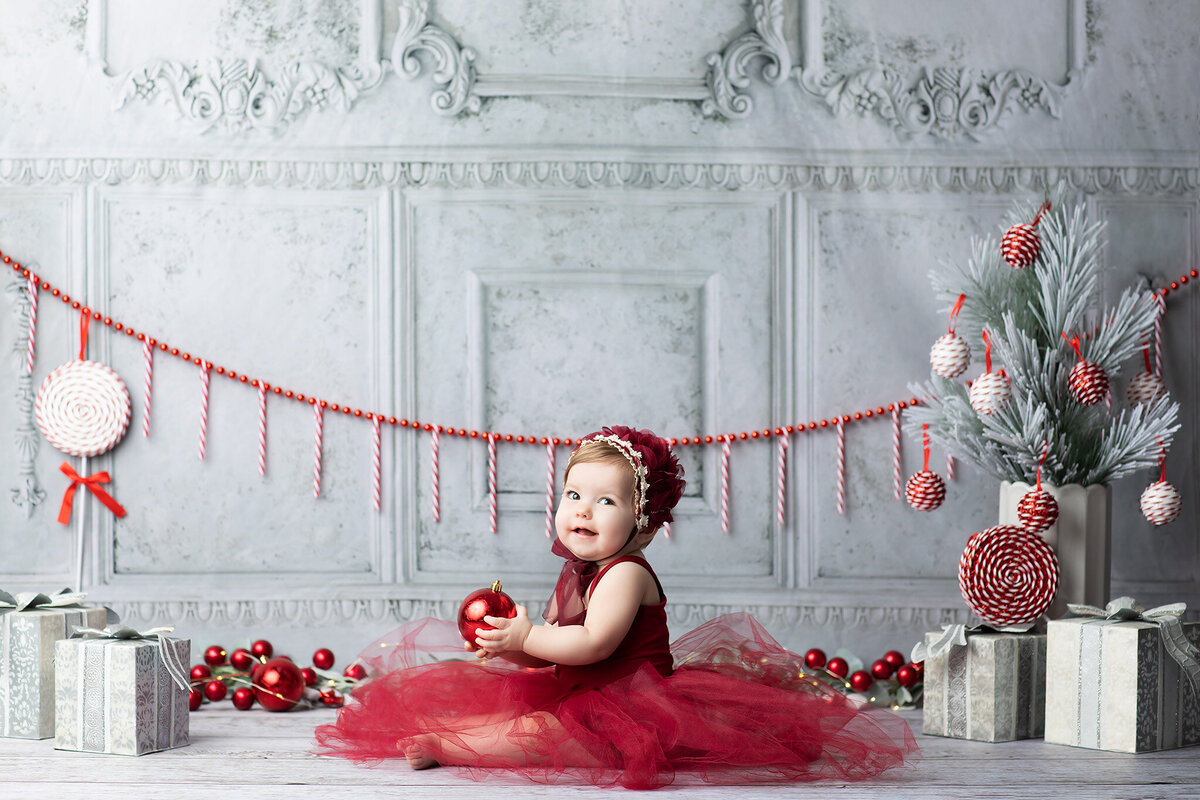 Smiling baby girl in a red tulle dress holding an ornament, surrounded by Christmas garlands, gifts, and peppermint candy decorations, captured by a Dallas milestone photographer.