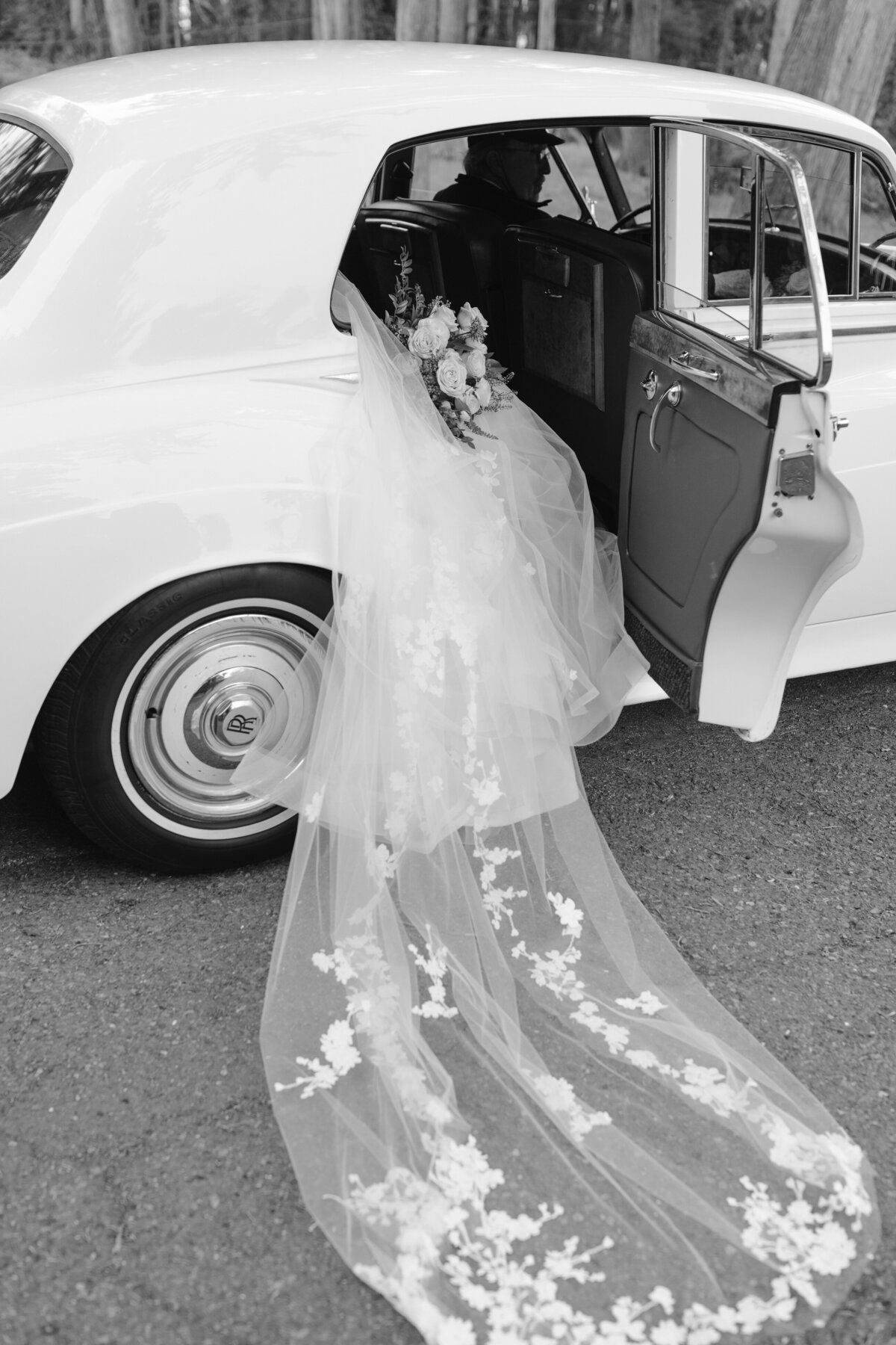 Bride's detailed veil and flowers flowing out of white vintage getaway car