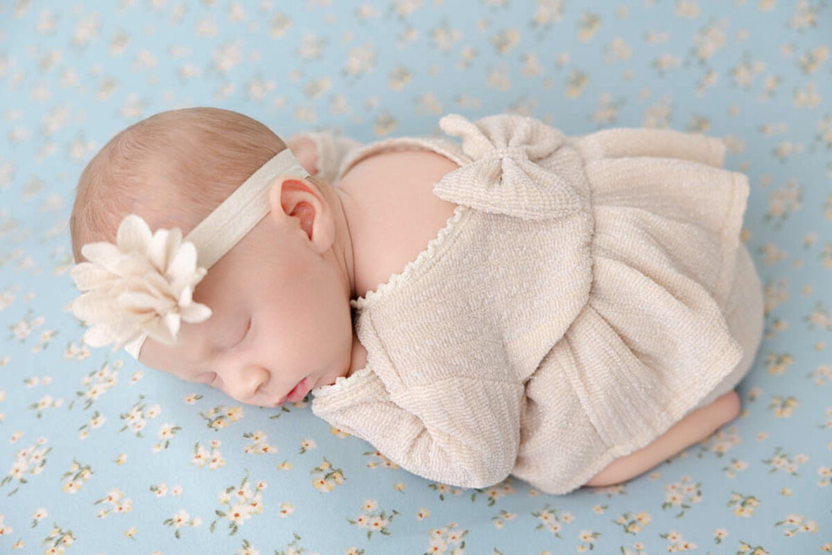 Baby girl newborn posed on a blue flowered blanket and wearing a champagne colored dress with a bow on her back and a cream-colored headband.
